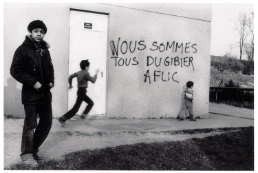 Photographie de Amadou Gaye dans la cité de transit de la Butte Rouge Chatenay-Malabry avec un graffiti "Nous sommes tous du gibier à flics" 